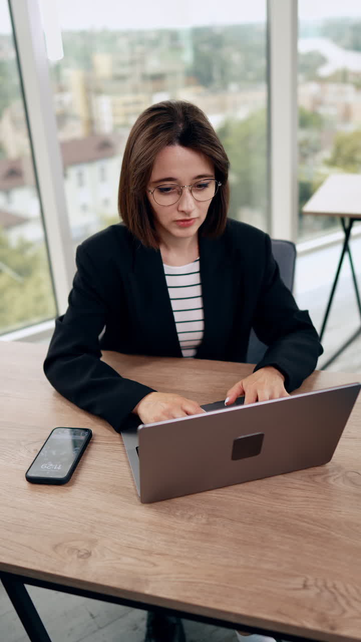 Good-looking young woman wearing glasses typing on her laptop. Business lady working in the office. Top view. Vertical video.