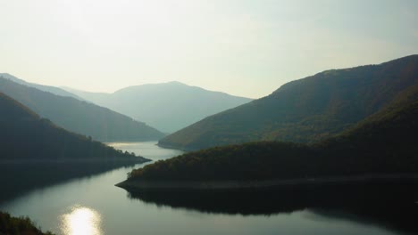 Aerial-view-of-huge-river-surrounded-by-forest-mountains,-sunny