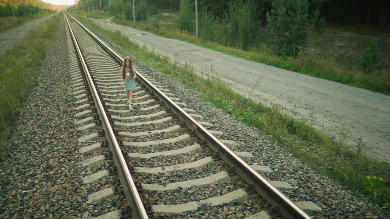 Little girl walking along railway track in countryside setting, surrounded by gravel and wild greenery, wearing denim dress and striped shirt with long hair down