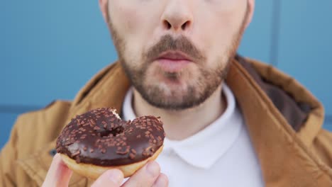 man with a mustache and a beard eats a chocolate doughnut against a blue background