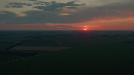 vibrant red sun dips below distant horizon illuminating towering dusk cloud with glowing flare while evening sky transitions from deep orange to muted blue tones over expansive open fields