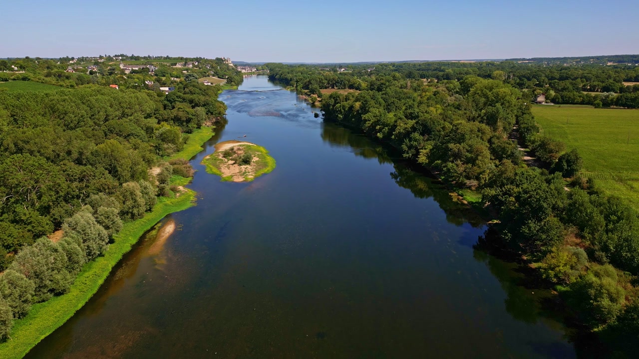 Aerial drone shot over the Vienne River with a small island, tree-lined banks, forest, and the town of Chinon visible in the distance.