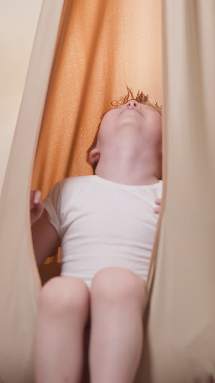 Relaxed little boy swaying in beige hammock hanging in gym. Happy toddler in white t-shirt enjoys indulging after intensive training slow motion