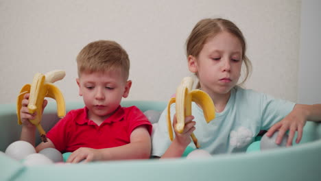 cute kids sitting together in pastel ball pit eating bananas happily, boy in red shirt holding banana while girl in blue shirt bites into hers, playful mood captured with bright soft light