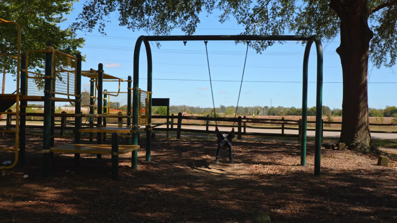 la cámara graciosamente empuja en un niño pequeño balanceándose un poco erráticamente, encarnando la alegría pura y la libertad, en un columpio del patio de recreo mientras un tren pasa en el fondo