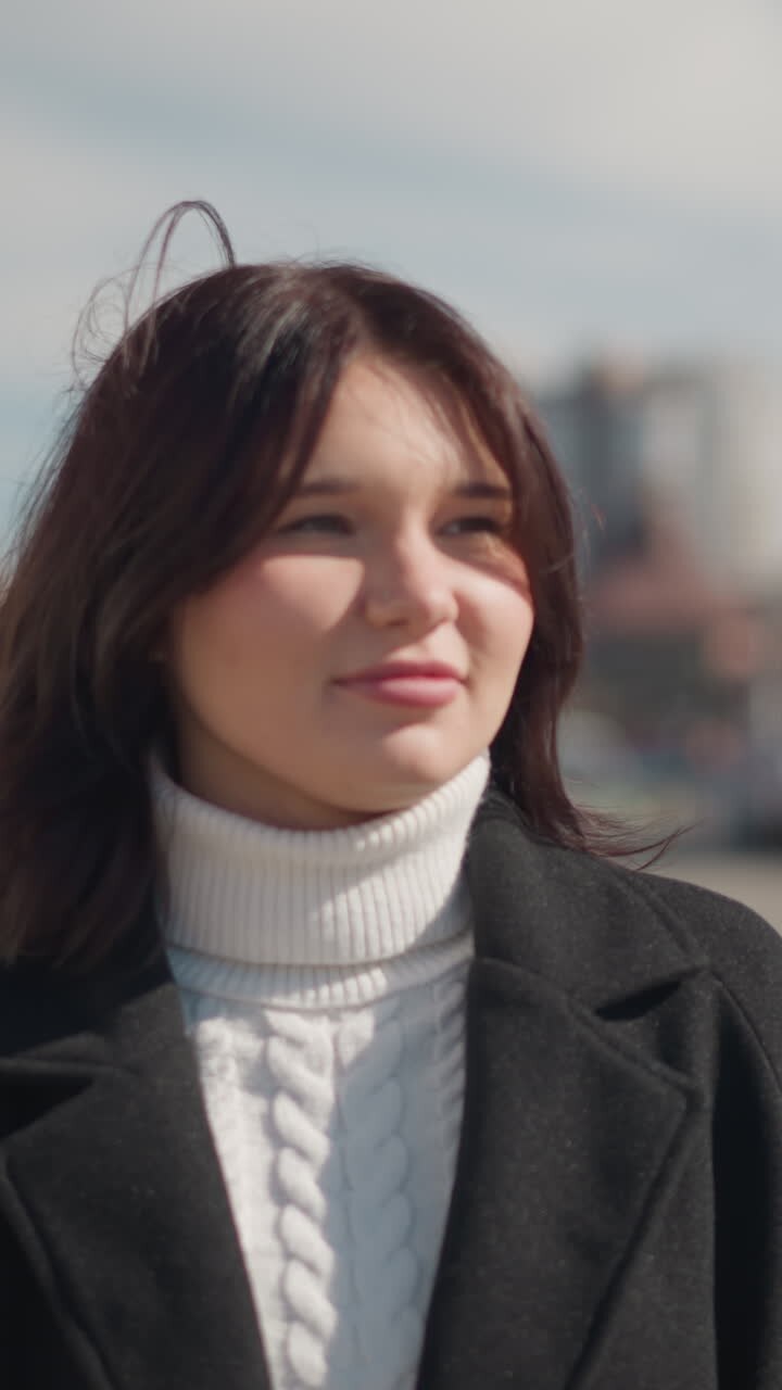 Close-up of a smiling woman in winter outfit standing outdoors on a sunny day in urban setting, with blurred background of city buildings
