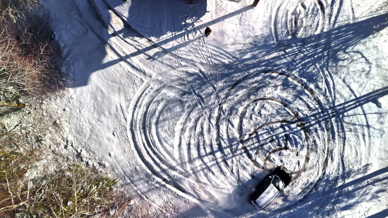 A car spins donuts in snow until it starts smoking heavily; nearby people rush to fan away the fumes in a chaotic winter mishap - top down view