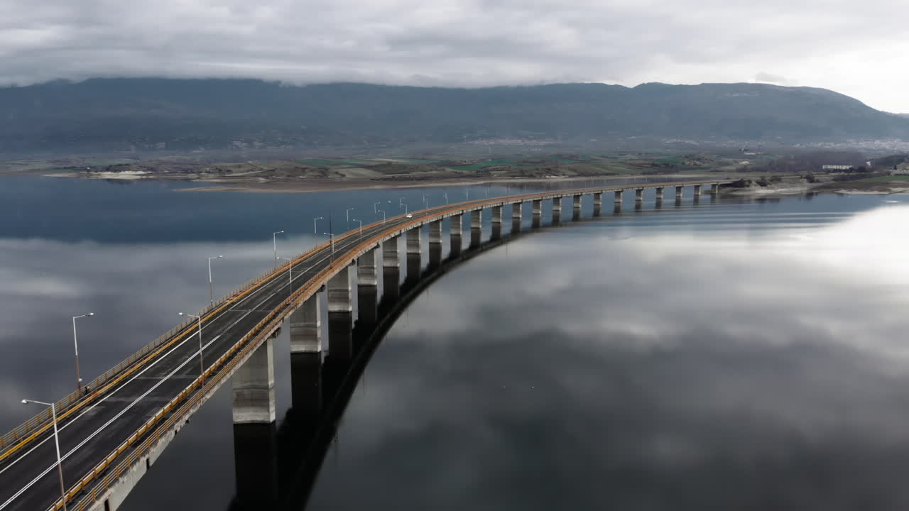 wide aerial View of High Bridge of Servia at lake polyfytos Kozani greece while tracking following cars , we see the reflection of the clouds at the lake and the bridge at the middle of the frame