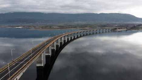 wide aerial view of high bridge of servia at lake polyfytos kozani greece while tracking following cars , we see the reflection of the clouds at the lake and the bridge at the middle of the frame