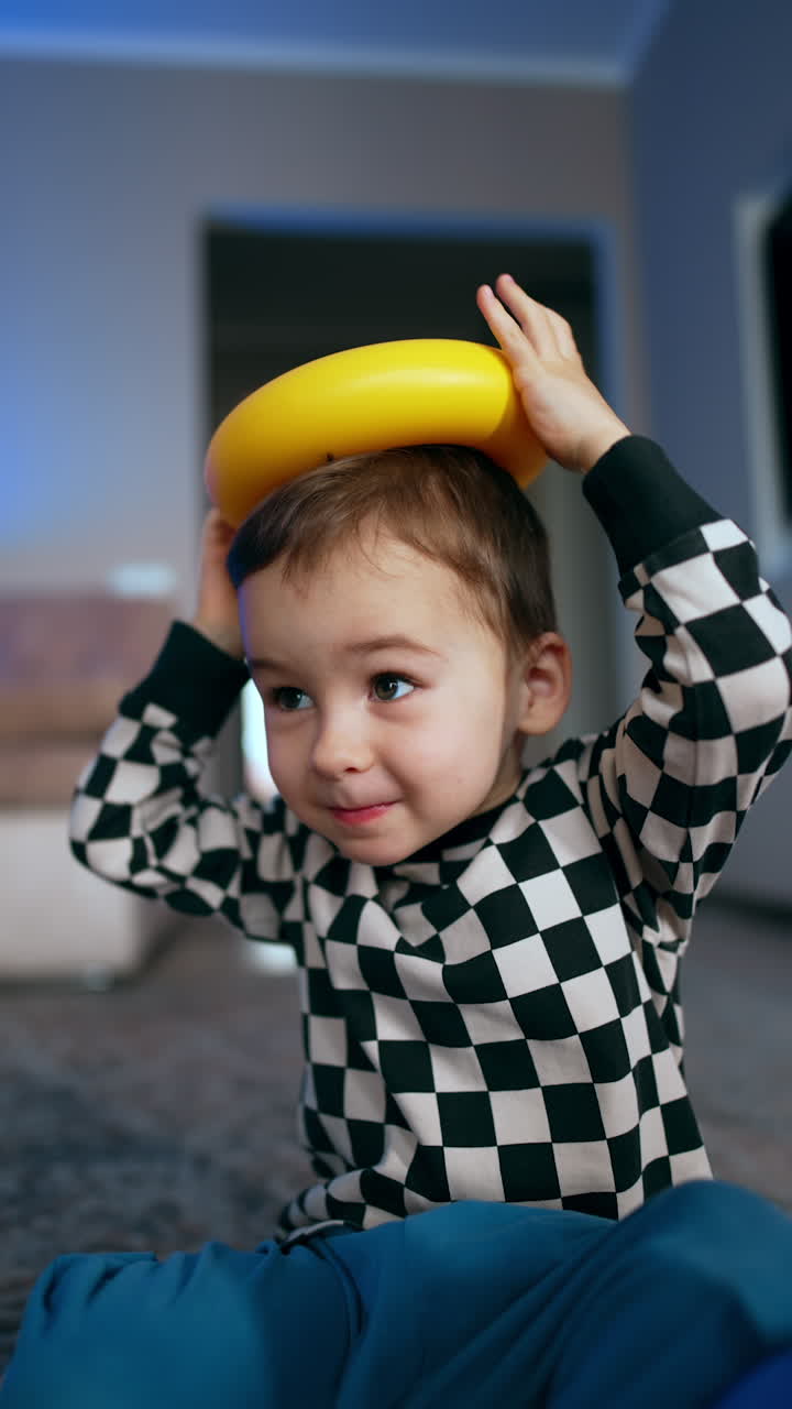 Beautiful Caucasian child puts a yellow piece of toy pyramid on his head. Happy baby playing with toys at home. Vertical video.