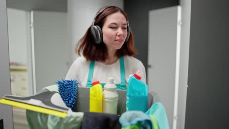 una confiada dama de limpieza morena con auriculares inalámbricos negros una camiseta blanca y un delantal azul camina a lo largo del corredor del apartamento y lleva un lavabo de plástico gris con detergentes y herramientas de limpeza en un apartamento moderno