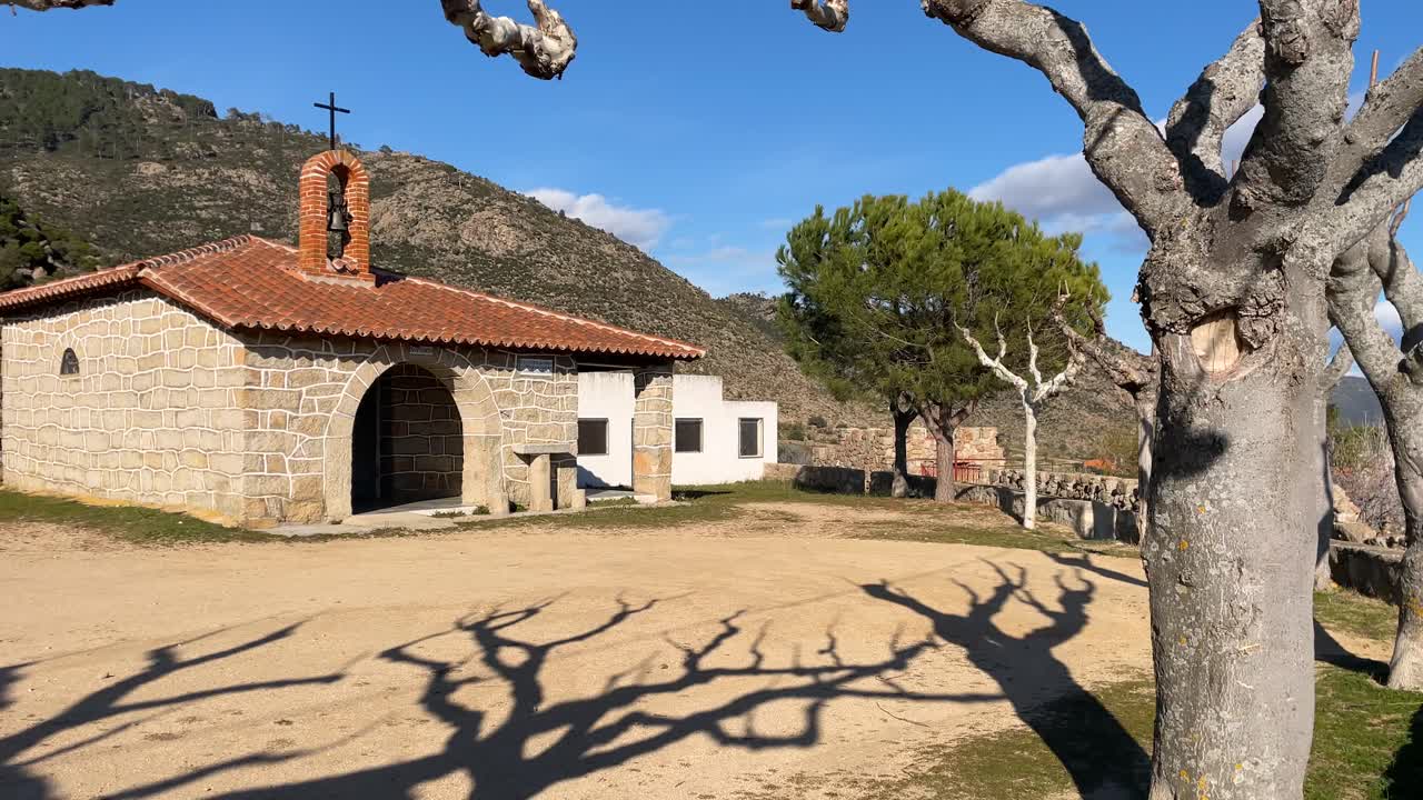 vemos con un giro de la cámara la ermita de san marcos en el barraco avila está hecha de piedra y en el techo está el pequeño campanario tiene una plaza de tierra con árboles podados en invierno españa