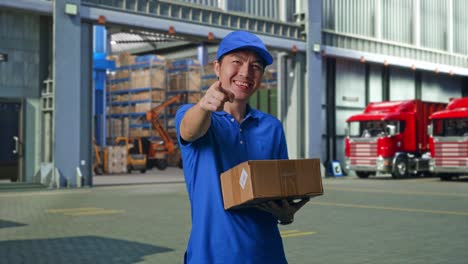 asian male courier in blue uniform smiling and making honest gesture while delivering a carton, outside of logistics distributions warehouse