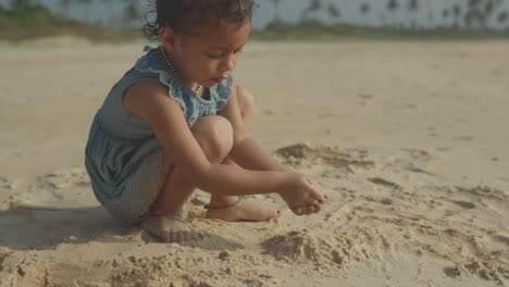 Young-brown-skinned-toddler-plays-in-sand-at-beach-patting-pile