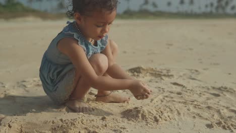 young brown skinned toddler plays in sand at beach patting pile