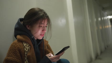 a close-up shot of a girl in a brown coat, sitting bent over in an underpass tunnel, scrolling through her phone with a heavy-hearted expression. moment of sorrow and reflection