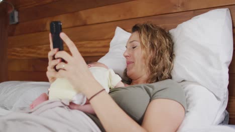 video de una madre caucásica feliz acostada en la cama con un bebé recién nacido durmiendo y usando un teléfono inteligente