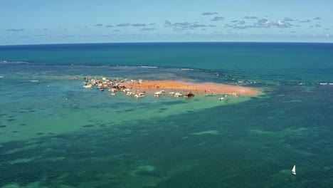 Dolly-in-aerial-wide-shot-revealing-the-Red-Sand-Island-in-the-tropical-capital-Joao-Pessoa,-Paraiba,-Brazil-with-dozens-of-tourist-boats-docked-and-people-enjoying-the-ocean-on-a-warm-summer-day