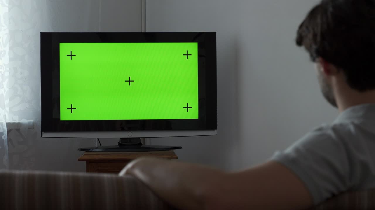 소파에 앉아서 녹색 스크린으로 tv를 보고 있는 젊은 남자