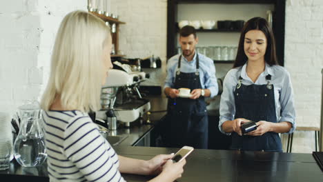 parte trasera de la mujer rubia que paga con su smartphone en el bar mientras compra un café y el camarero lo sirve