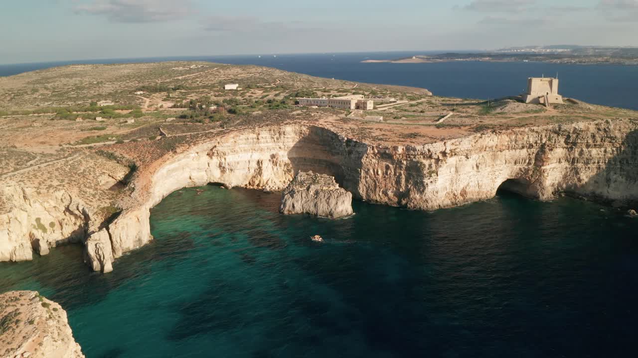 Aerial drone view of Malta's Blue Lagoon, with the drone circling boats and revealing the stunning rugged rock formations and coastal caves adorning Como Island's picturesque shoreline LuPa Creative
