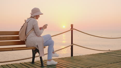 a stylish girl in a pink hat sits on a bench on a sea pier and reads a text message on the phone the