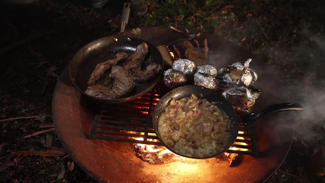 A campfire cooking feast with steak stew and potatoes in foil being roasted on a fire.