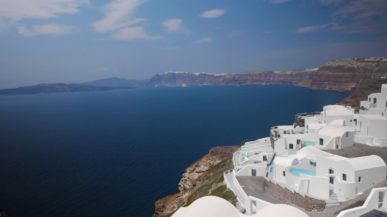 toma amplia de mano de un hotel en un acantilado con edificios blancos y una vista de los acantilados de santorini hasta la ciudad de thira