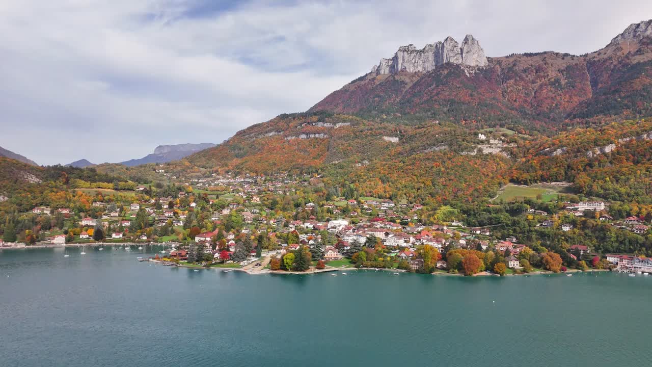 Aerial view of Chateau de Duingt, France, featuring a lakeside village, turquoise water, and colorful autumn foliage set against dramatic mountain peaks and a peaceful rural landscape