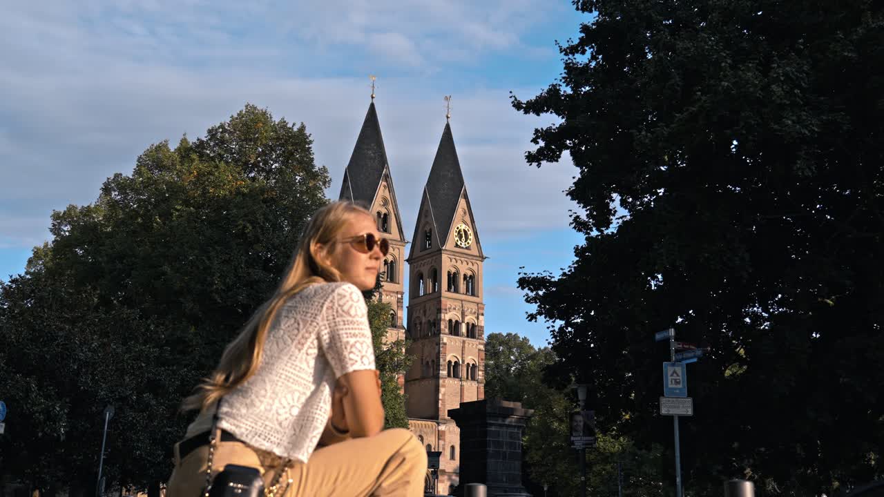 Blonde woman admiring the historic Basilica of St. Castor in Koblenz, Germany, with its twin Romanesque towers and dark pyramidal roofs dominating the background