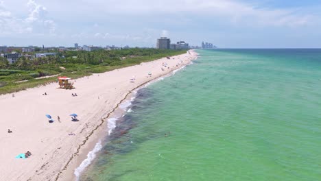 drone flying over miami beach, people sunbathing and swimming in ocean, iconic lifeguard tower and city skyline in distance, florida. aerial