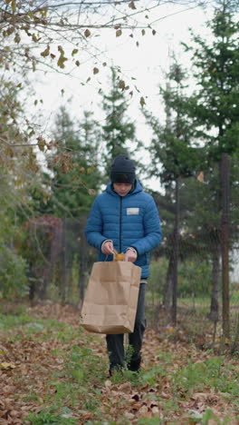 joven en chaqueta azul y gorra se inclina para recoger la hoja amarilla del suelo, colocándola en una bolsa de papel en medio de la vegetación al aire libre vibrante y las hojas de otoño caídas, al lado de una valla rústica