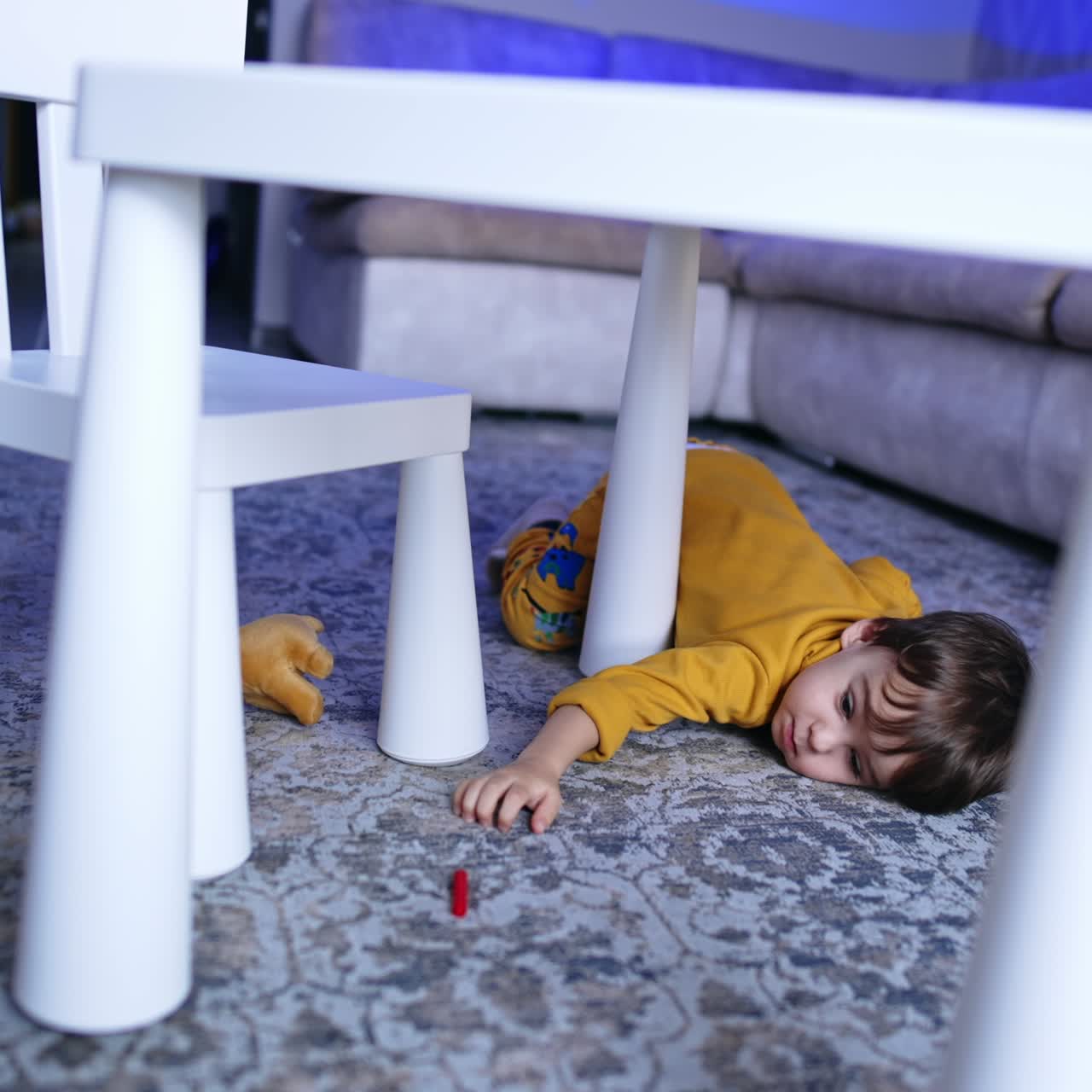 Small kid lies on the floor near his desk. Toddler tries to get the cap from a felt pen. Boy stands up and walks to the seat