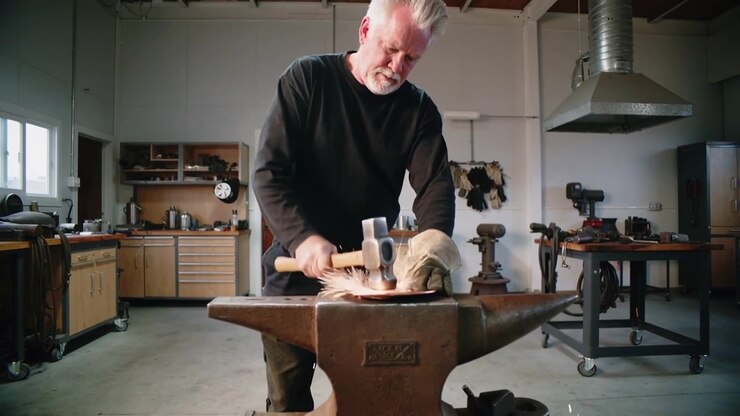 Placing disc on anvil, craftsman in black shirt hammering at forge with hammer, gloved shaping disc