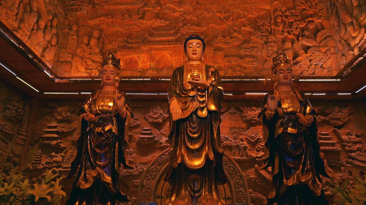 Vietnam, Ninh Binh'deki Bai Dinh Pagoda kompleksinde, bir tapınağın içinde, özenle oyulmuş turuncu bir duvarın önünde üç görkemli altın Buda heykeli duruyor.
