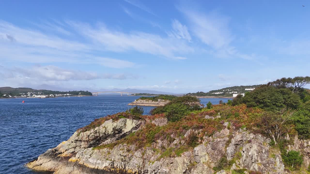 Smooth daytime camera pan reveals rugged rocky coastline, green vegetation, and calm blue waters