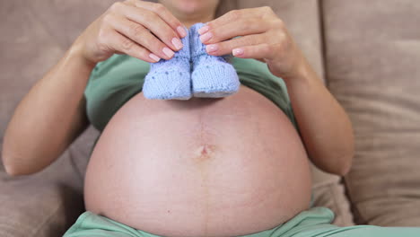 beautiful blue knitted socks in hands of a pregnant woman. big belly of a future mother laying on a sofa at home. close-up. happy pregnancy.