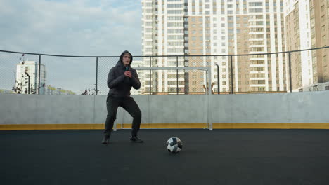 sportsman performing squats with hands clasped on outdoor sports ground preparing for exercise, soccer ball in foreground, urban high-rise buildings, and goal post