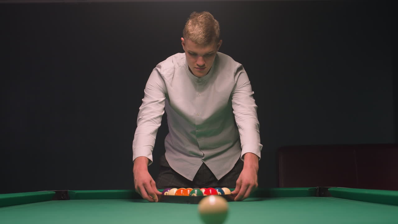 primer plano de un joven en camisa intensamente enfocado en la mesa de billar mientras se prepara para romper. sus manos se mueven sobre un estante de bolas de colores en fieltro verde vibrante, luz suave contra un fondo oscuro