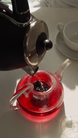 pouring hot water into teapot on table closeup. brewing hibiscus tea with flow from electric kettle for breakfast in shady kitchen. healthy beverage