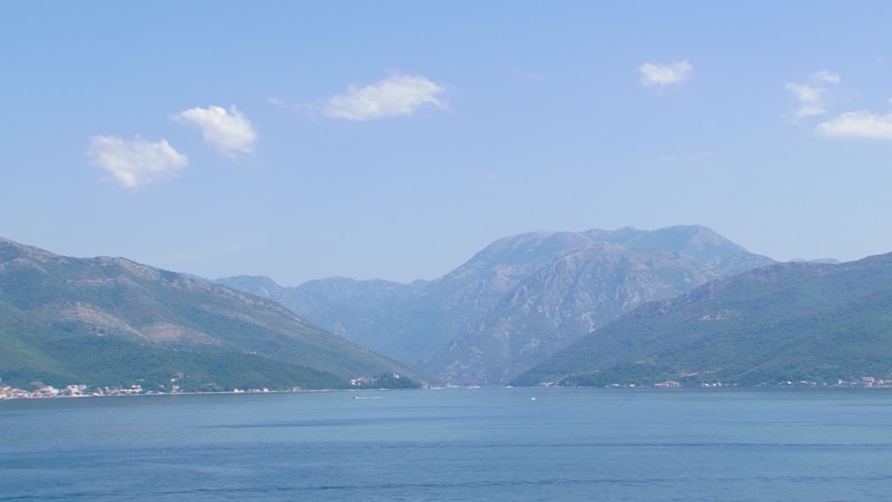 보카 코토르스카 베이, 깨끗한 아드리아 해수를 항해하는 보트, 푸른 하늘에 약간의 구름이 있는 산이 뒤에 있습니다.