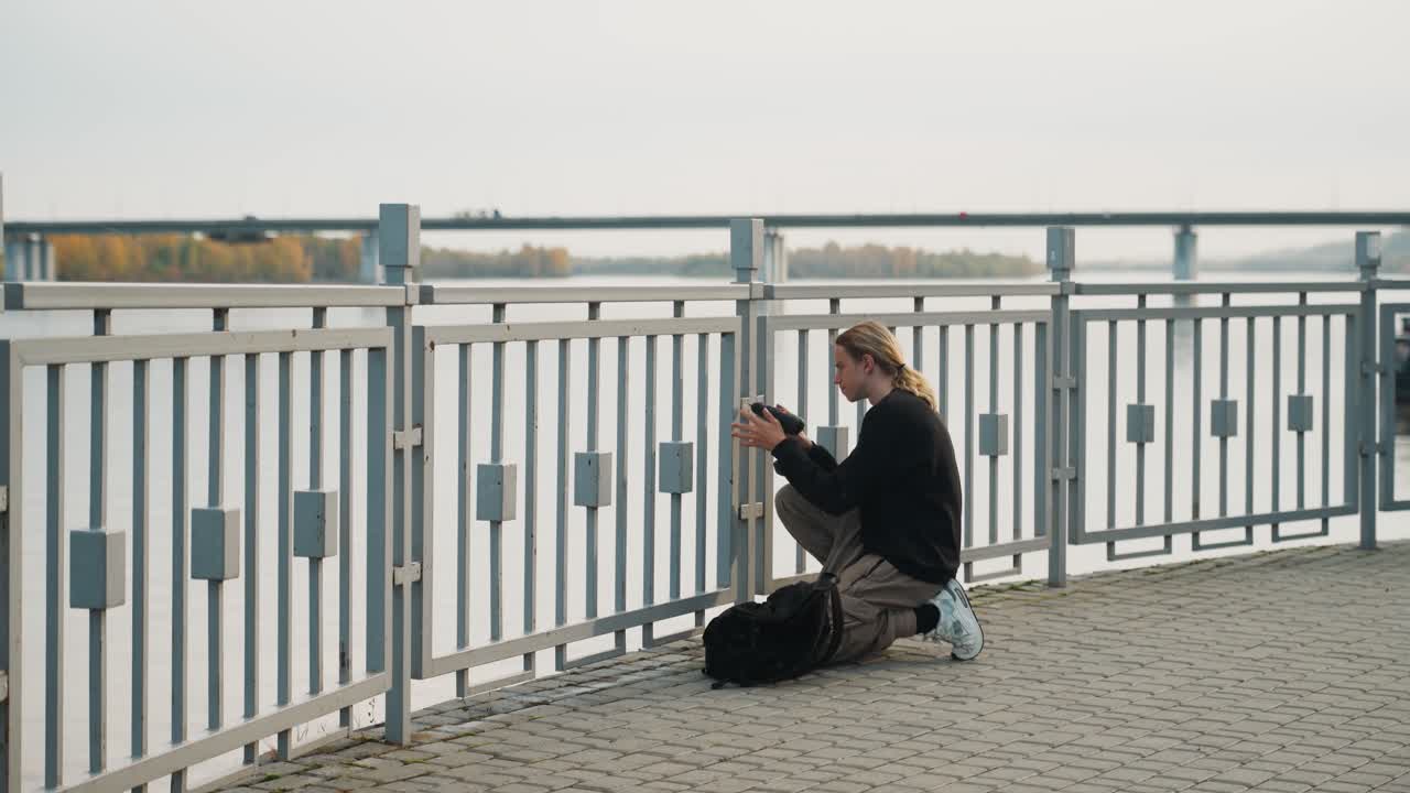 Fotograf gennemser billeder. Knælende fotograf kalibrerer enhed nær flod. Fokuseret ung fotograf dukker sig ved vandet, gennemser billeder på sin enhed i rolige efterårsomgivelser.
