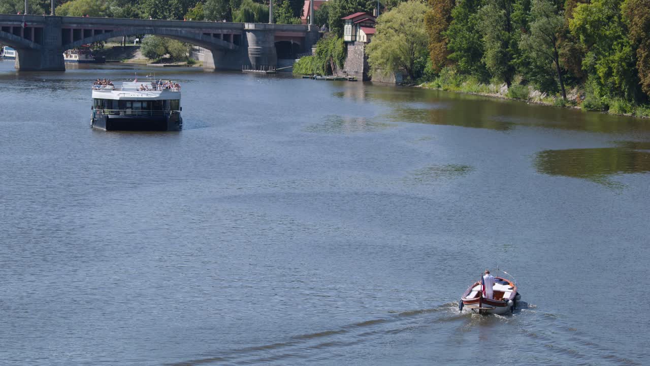Two boats travel on Vltava River, Prague, under bridges, with lush riverside trees, daylight