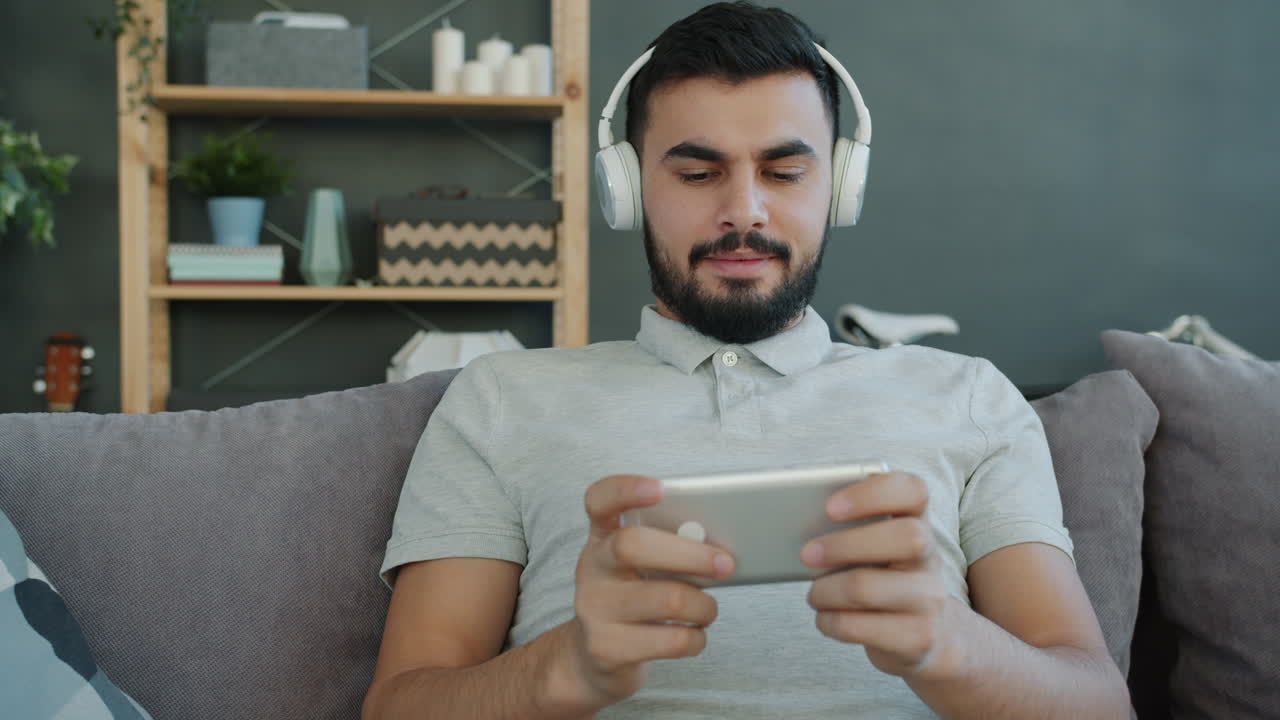 Man Relaxing on Couch, Playing Games on Smartphone with Headphones