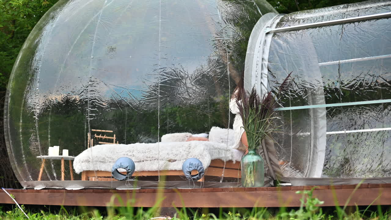 Tenda a bolla trasparente in un campeggio di lusso, foresta lussureggiante intorno e interni. Donna con una tazza che guarda fuori