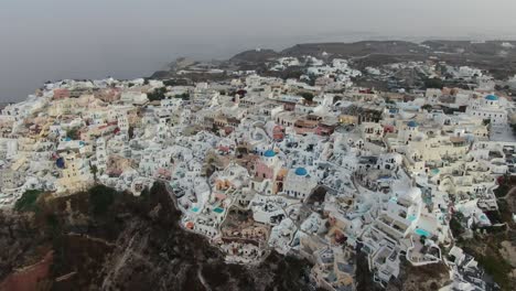 drone view in greece flying over santorini with oia town white houses and blue roofs on a cliff next to the mediterranean sea at sunrise