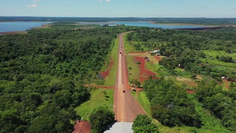 Drone-camera-flying-backwards-revealing-a-tropical-road-in-Misiones,-Argentina-where-you-can-see-the-jungle-and-a-large-lake-seen-from-above