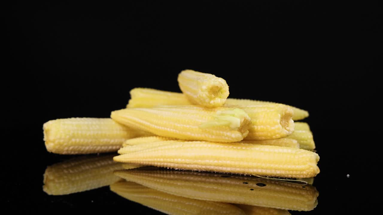 Baby corn rotates on a glossy black surface, highlighting texture and color under bright lighting