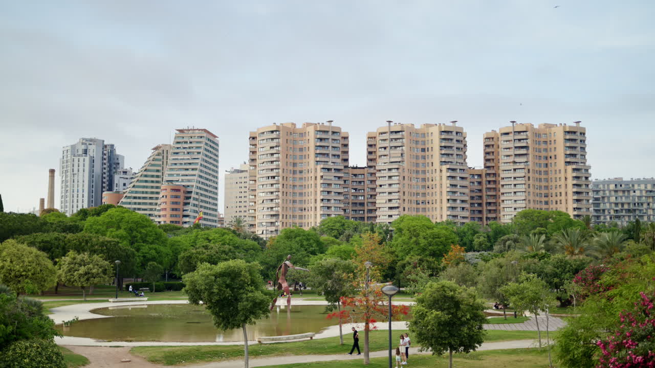 Valencia, Spain - May 28, 2025: View of the Jardi del Turia showing a pond and sculpture framed by lush trees, with modern residential towers on the skyline under bright overcast light