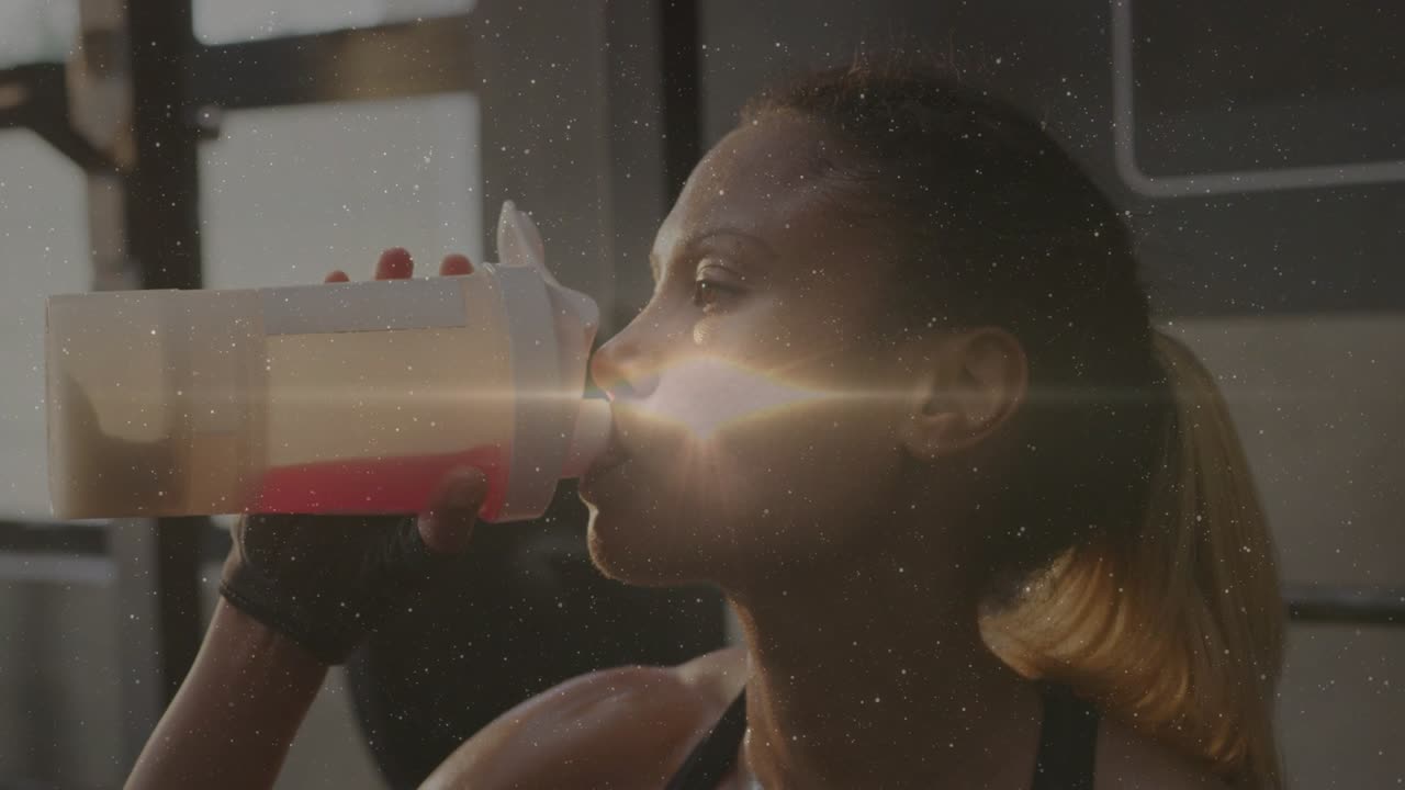 Female athlete inspecting shaker lid then sipping red drink in gym, pausing and breathing